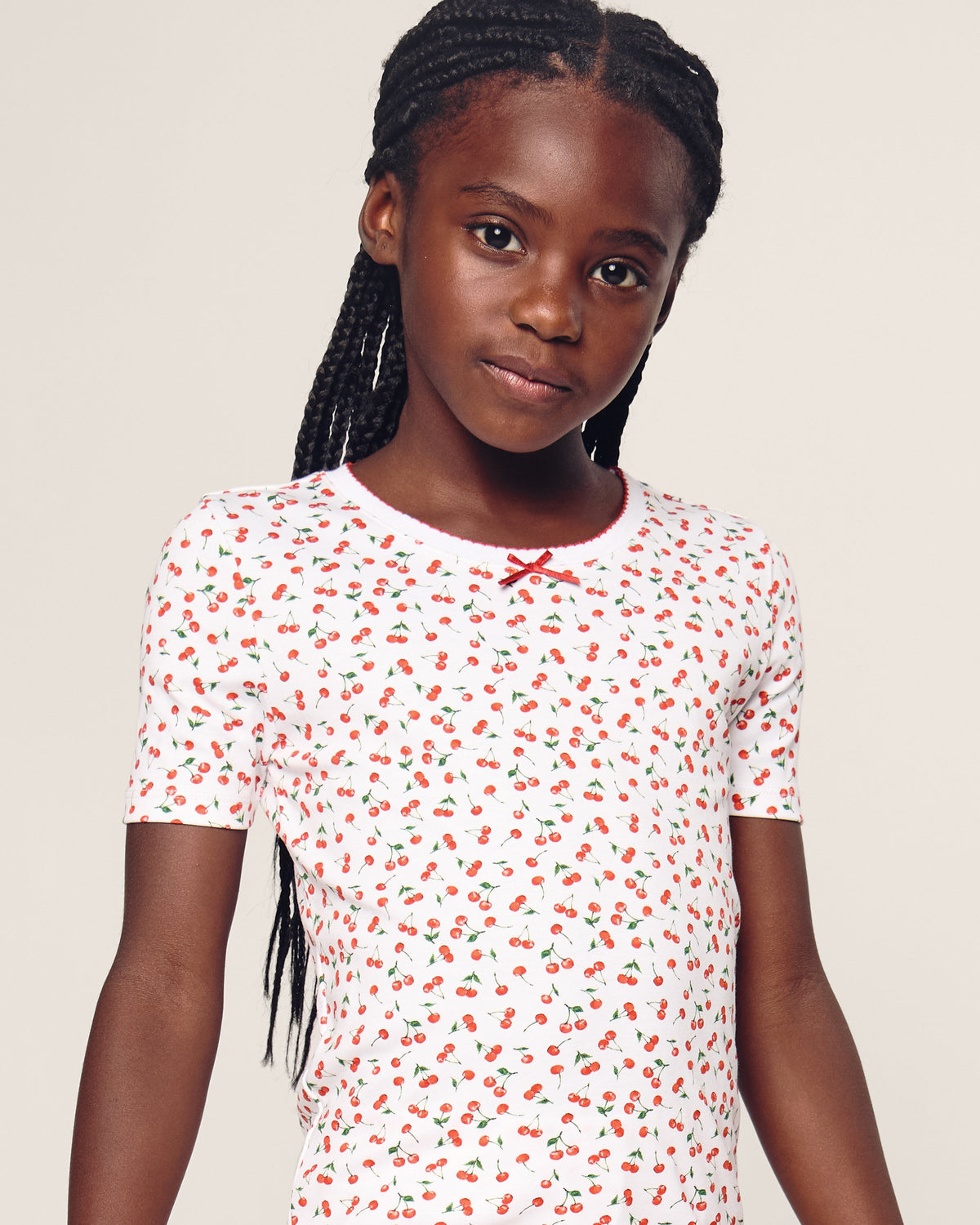 A young girl with braided hair stands against a plain background, wearing Petite Plumes Kids Pima Snug Fit Short Set in Cherry Orchard. The white Pima Cotton T-shirt features a red cherry pattern and a small bow at the neckline, adding to its stylish silhouette.