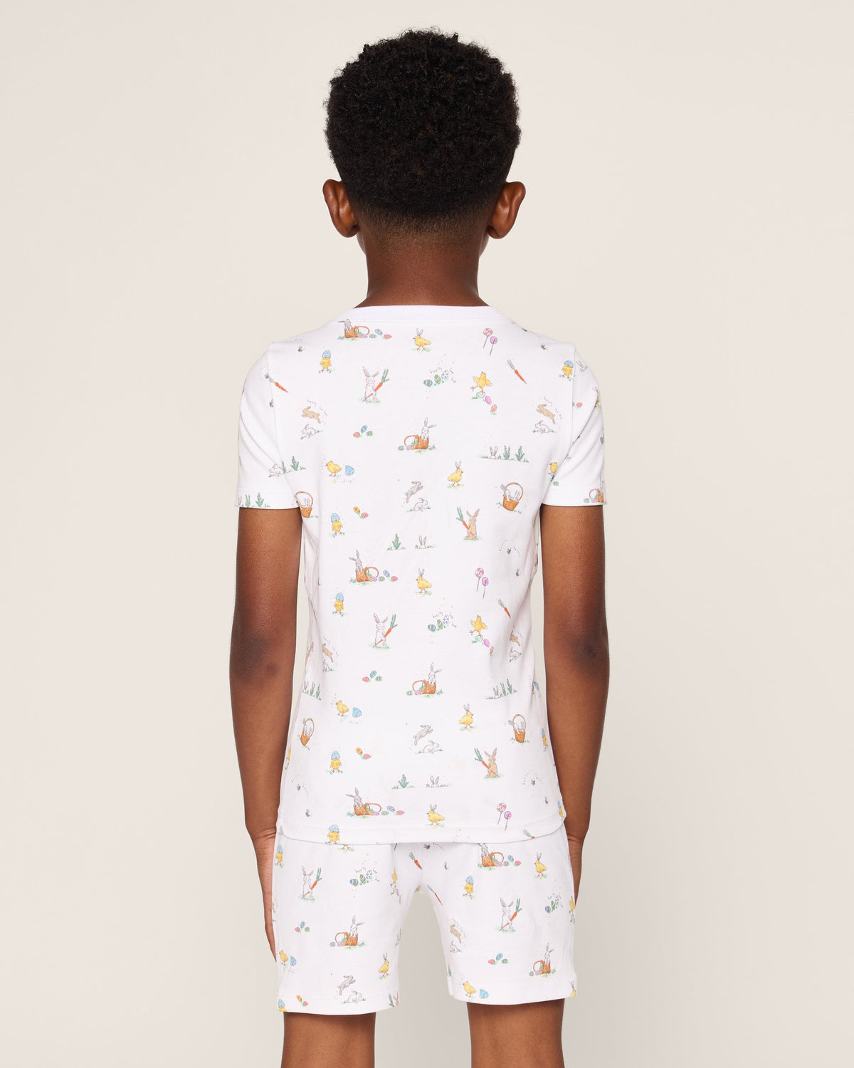 A child faces away in a Petite Plume Kids Pima Snug Fit Short Set in Easter Frolic prints. The white sleepwear, made from soft Pima Cotton, features colorful animal and floral designs against a plain beige background.