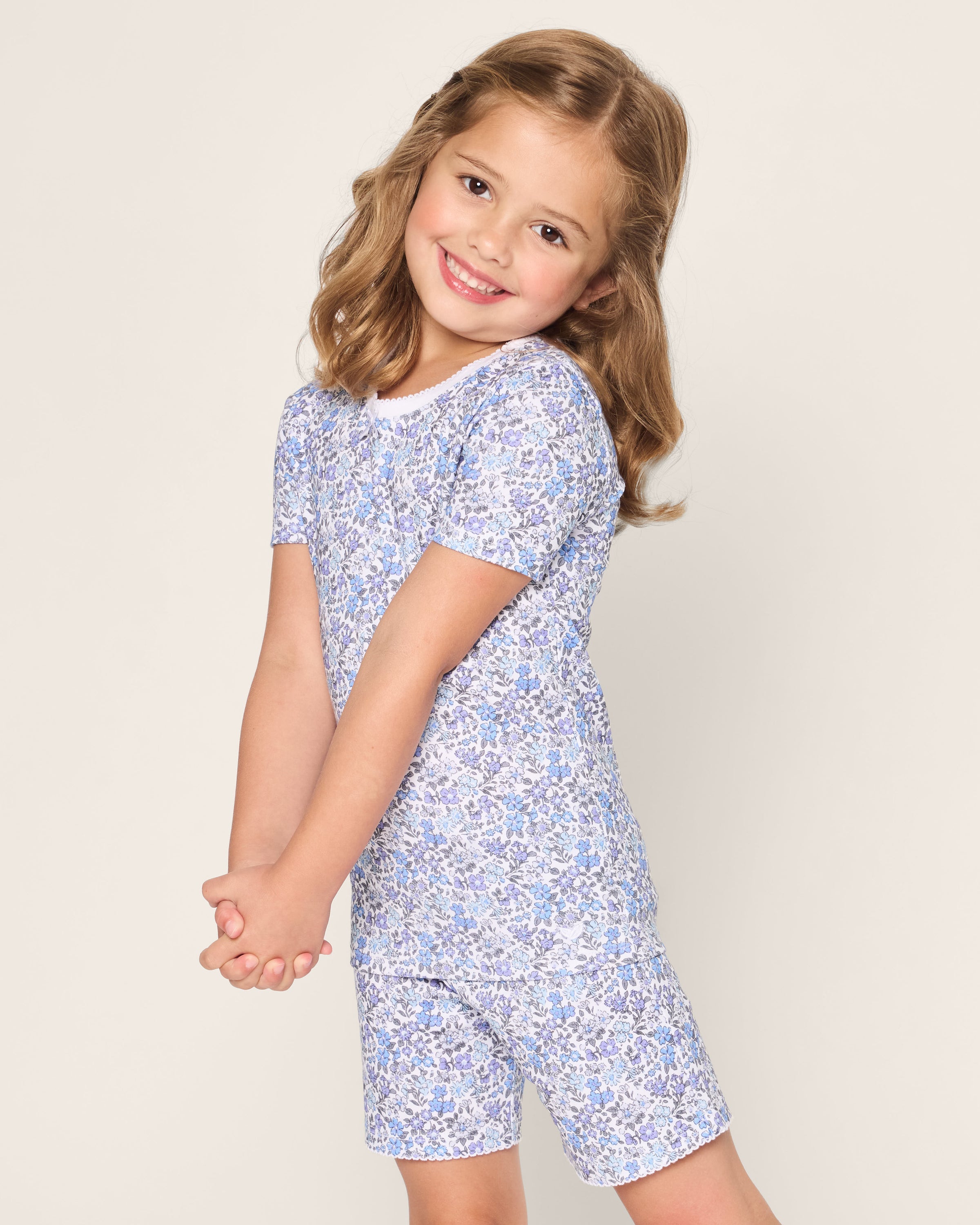 A young girl with shoulder-length light brown hair smiles, wearing Petite Plumes Kids Pima Snug Fit Short Set in Fleur dAzur. She stands against a plain background, tilting her head slightly and holding her hands together in front.