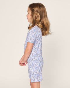 A young girl with light brown hair stands in profile against a beige backdrop, wearing Petite Plumes Kids Pima Snug Fit Short Set in Fleur dAzur, a vintage-inspired blue floral print outfit.