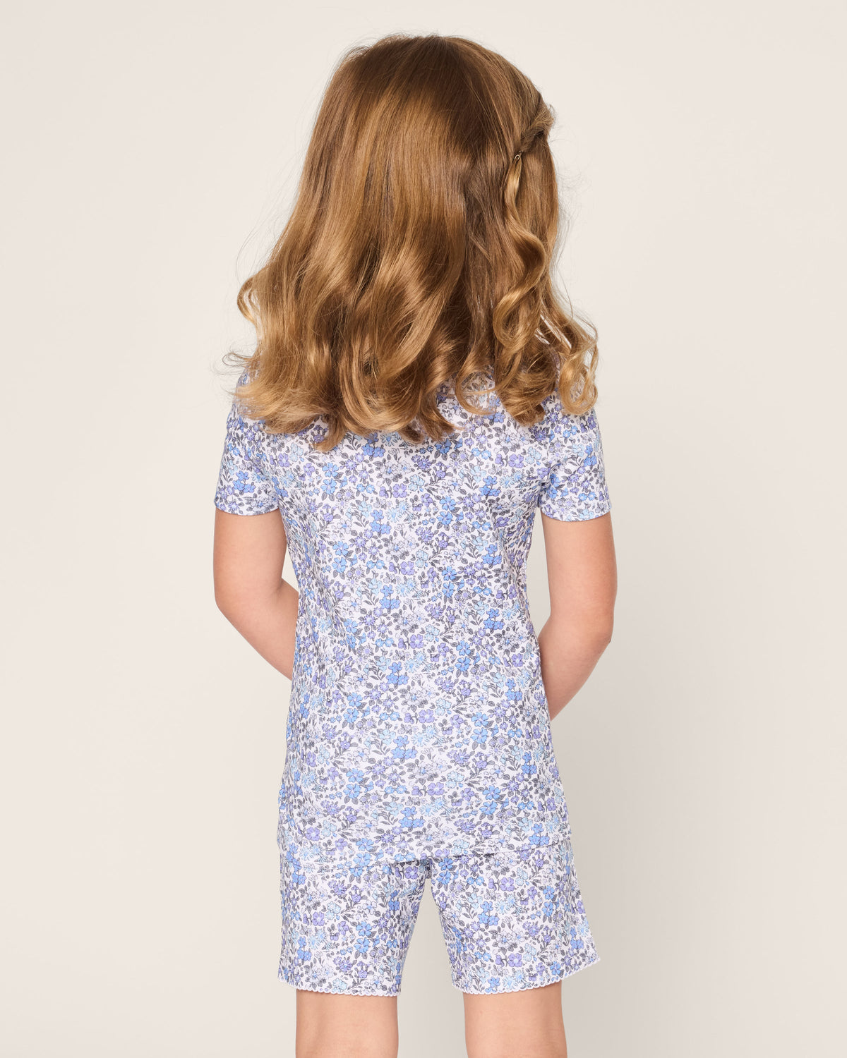 A young child with wavy brown hair wears Petite Plumes Kids Pima Snug Fit Short Set in Fleur dAzur, featuring a vintage-inspired blue floral pattern, as they stand facing away against a light background.