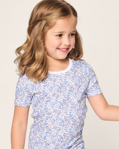 A young girl with wavy blonde hair smiles wearing Petite Plumes Kids Pima Snug Fit Short Set in Fleur dAzur, featuring a blue and white floral pattern in a vintage-inspired print. She gazes to the side against a plain background.