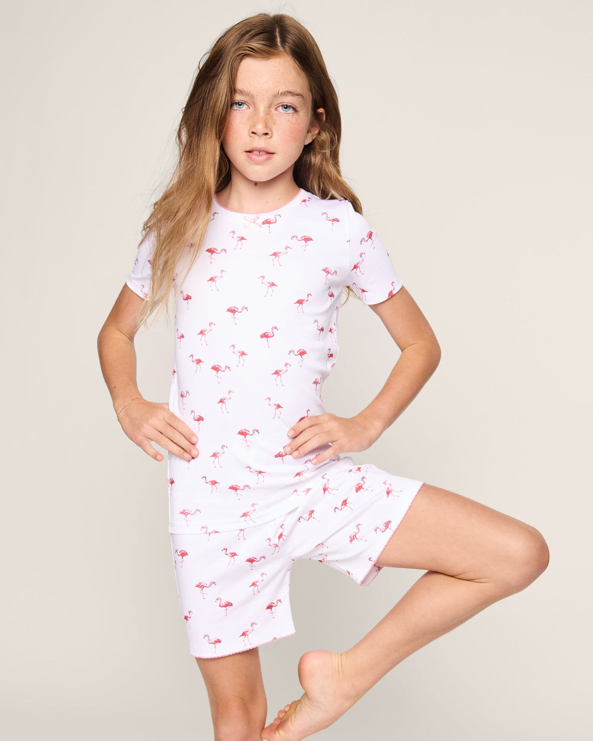 A young girl strikes a confident pose in Petite Plumes Kids Pima Snug Fit Short Set in Flamingo. The cozy white set, adorned with pink flamingos, complements her playful charm as she stands against a light background with one hand on her hip and her other leg bent.