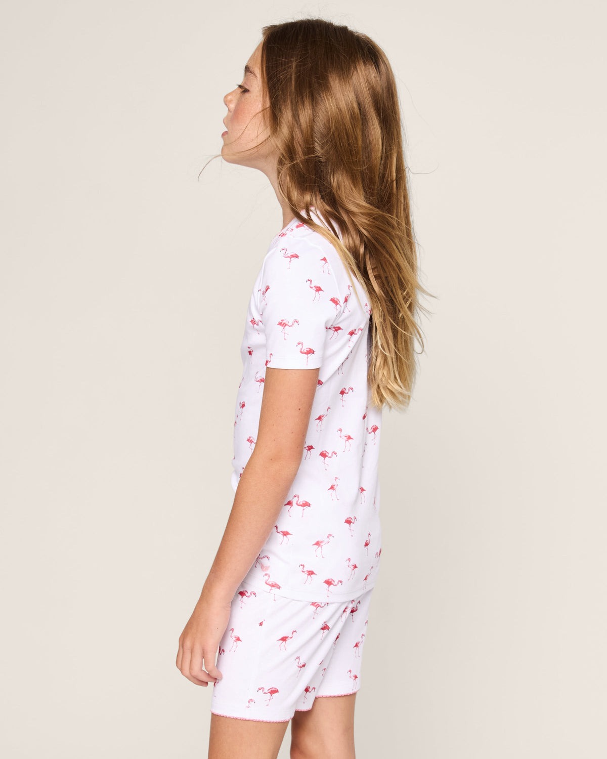 A young girl with long blonde hair stands sideways, looking upwards in her stylish Petite Plume Kids Pima Snug Fit Short Set in Flamingo. She wears a cozy white ensemble against a plain off-white background, making her playful attire pop.