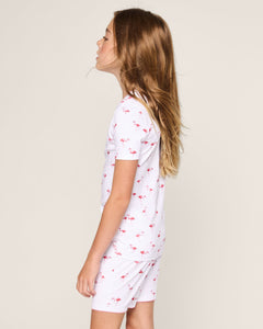 A young girl with long blonde hair stands sideways, looking upwards in her stylish Petite Plume Kids Pima Snug Fit Short Set in Flamingo. She wears a cozy white ensemble against a plain off-white background, making her playful attire pop.