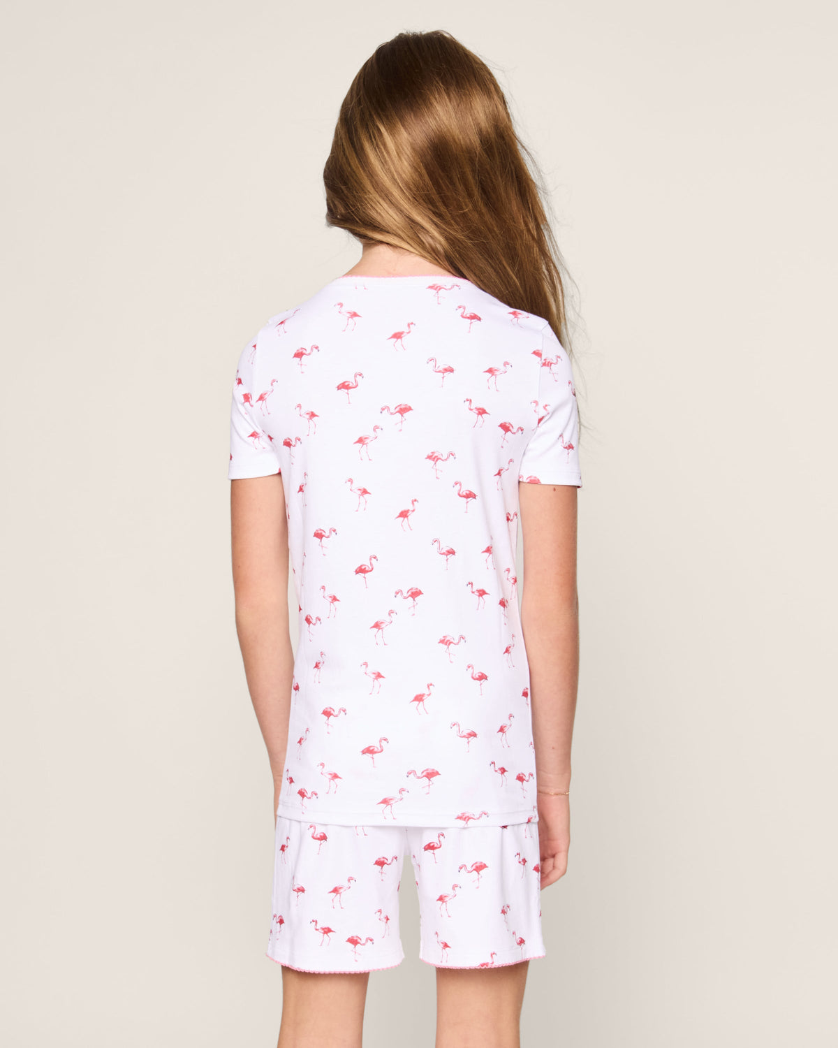 A child with long hair faces away from the camera, modeling Petite Plumes Kids Pima Snug Fit Short Set in Flamingo. This stylish pajama set features pink flamingo patterns on white, complementing an off-white backdrop.