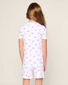 A child with long hair faces away from the camera, modeling Petite Plumes Kids Pima Snug Fit Short Set in Flamingo. This stylish pajama set features pink flamingo patterns on white, complementing an off-white backdrop.