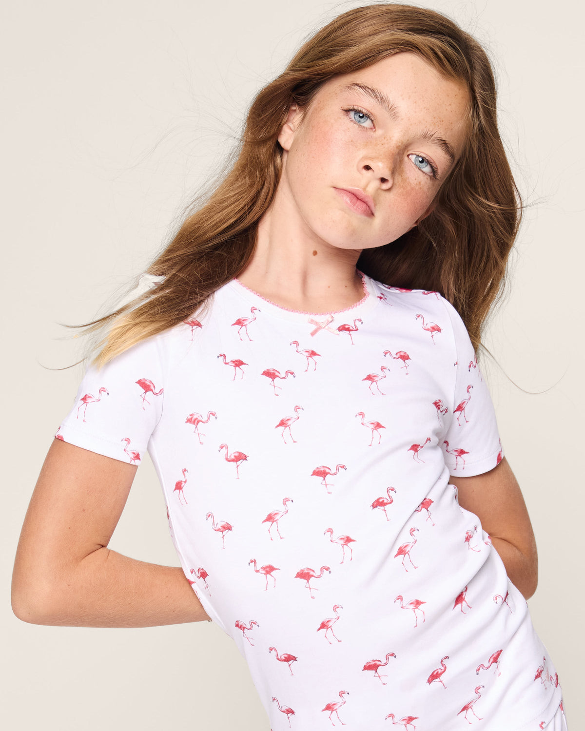 A young girl with long hair wears the Kids Pima Snug Fit Short Set in Flamingo by Petite Plume, featuring a stylish white t-shirt with pink flamingo prints. Made from soft Pima cotton sleepwear, she gazes neutrally at the camera against a plain light-colored background.