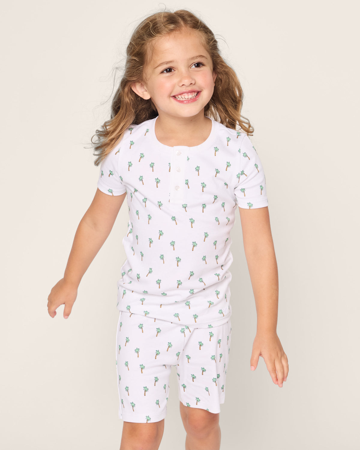 A young girl with long brown hair is wearing Petite Plumes Kids Pima Snug Fit Short Set in Palmier. She smiles, looking off to the side against a plain light background.
