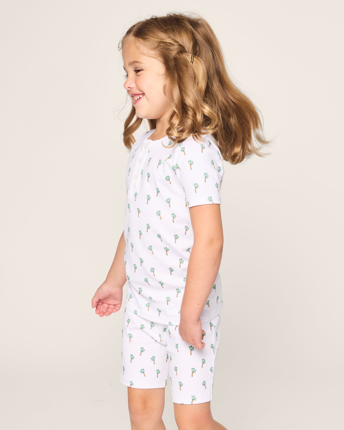 A young brown-haired girl stands smiling in Petite Plumes Kids Pima Snug Fit Short Set in Palmier, featuring matching white pajamas with small palm tree prints. The short sleeve top adds charm against the plain light background.
