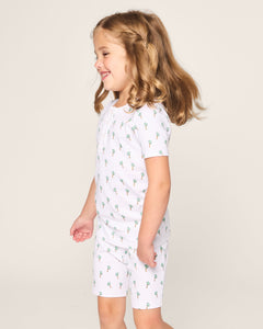 A young brown-haired girl stands smiling in Petite Plumes Kids Pima Snug Fit Short Set in Palmier, featuring matching white pajamas with small palm tree prints. The short sleeve top adds charm against the plain light background.