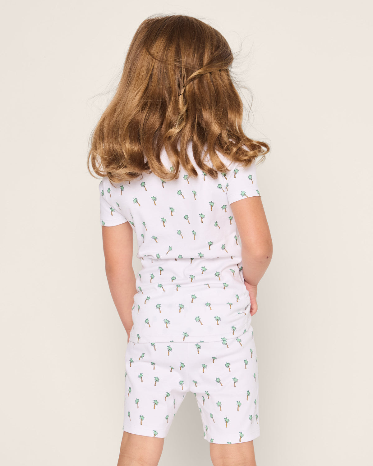 A child with medium brown hair is seen from the back, wearing Petite Plumes Kids Pima Snug Fit Short Set in Palmier. The white shorts and short sleeve top, made of soft pima cotton with small palm tree patterns, resemble sleepwear against the plain light background.