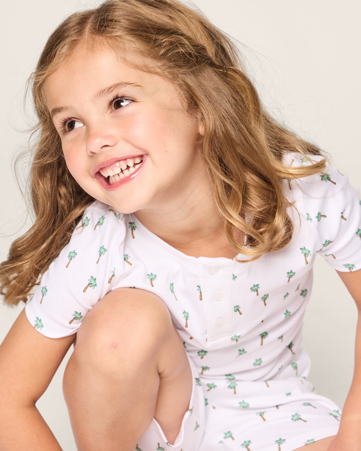 A cheerful girl with curly hair models Petite Plumes Kids Pima Snug Fit Short Set in Palmier, featuring a fun palm tree print and short sleeves, set against a light background.
