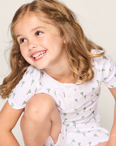A cheerful girl with curly hair models Petite Plumes Kids Pima Snug Fit Short Set in Palmier, featuring a fun palm tree print and short sleeves, set against a light background.