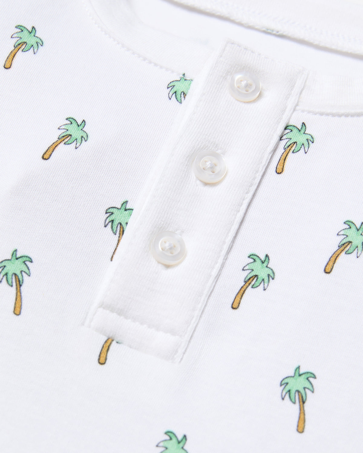 Close-up of the Kids Pima Snug Fit Short Set in Palmier by Petite Plume: white sleepwear shirt with a henley neck, four white buttons, soft Pima cotton, short sleeves, and small colorful palm trees scattered throughout.