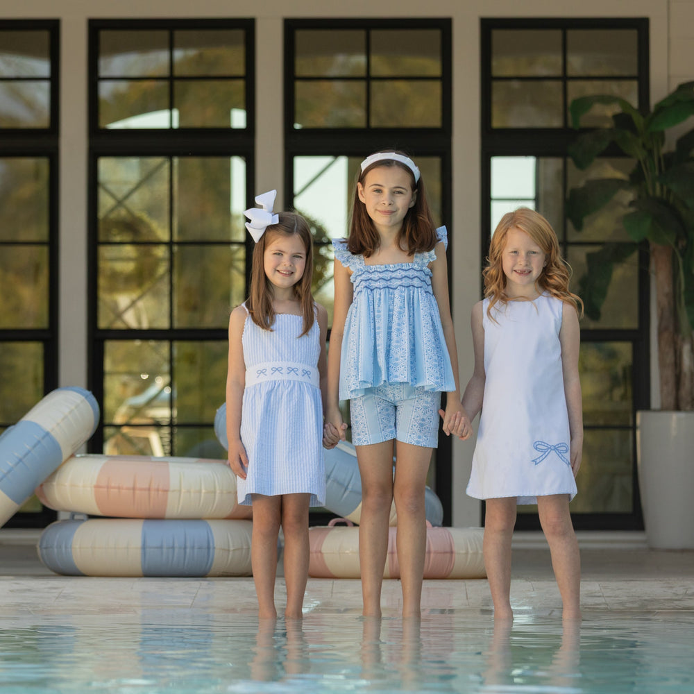 Three girls holding hands at a poolside entrance wearing coordinating blue and white summer dresses