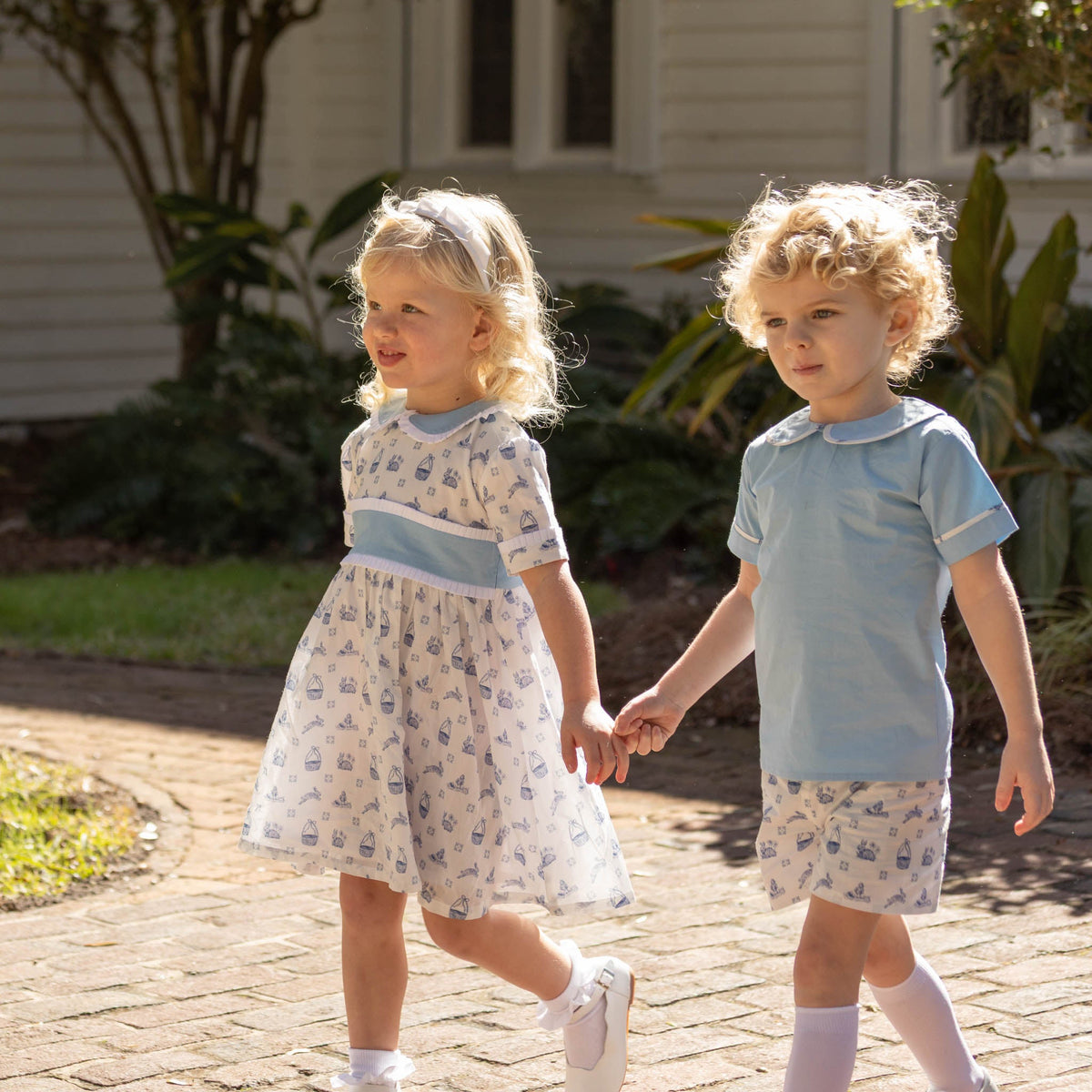 Two toddlers holding hands on a brick path, girl in white and blue dress, boy in light blue shirt with patterned shorts