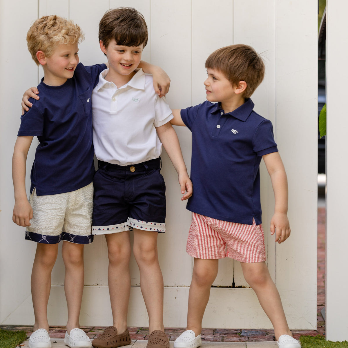 Three boys arm-in-arm by a white fence, one wearing Rhode Island Red seersucker Liam Shorts that hit above the knee