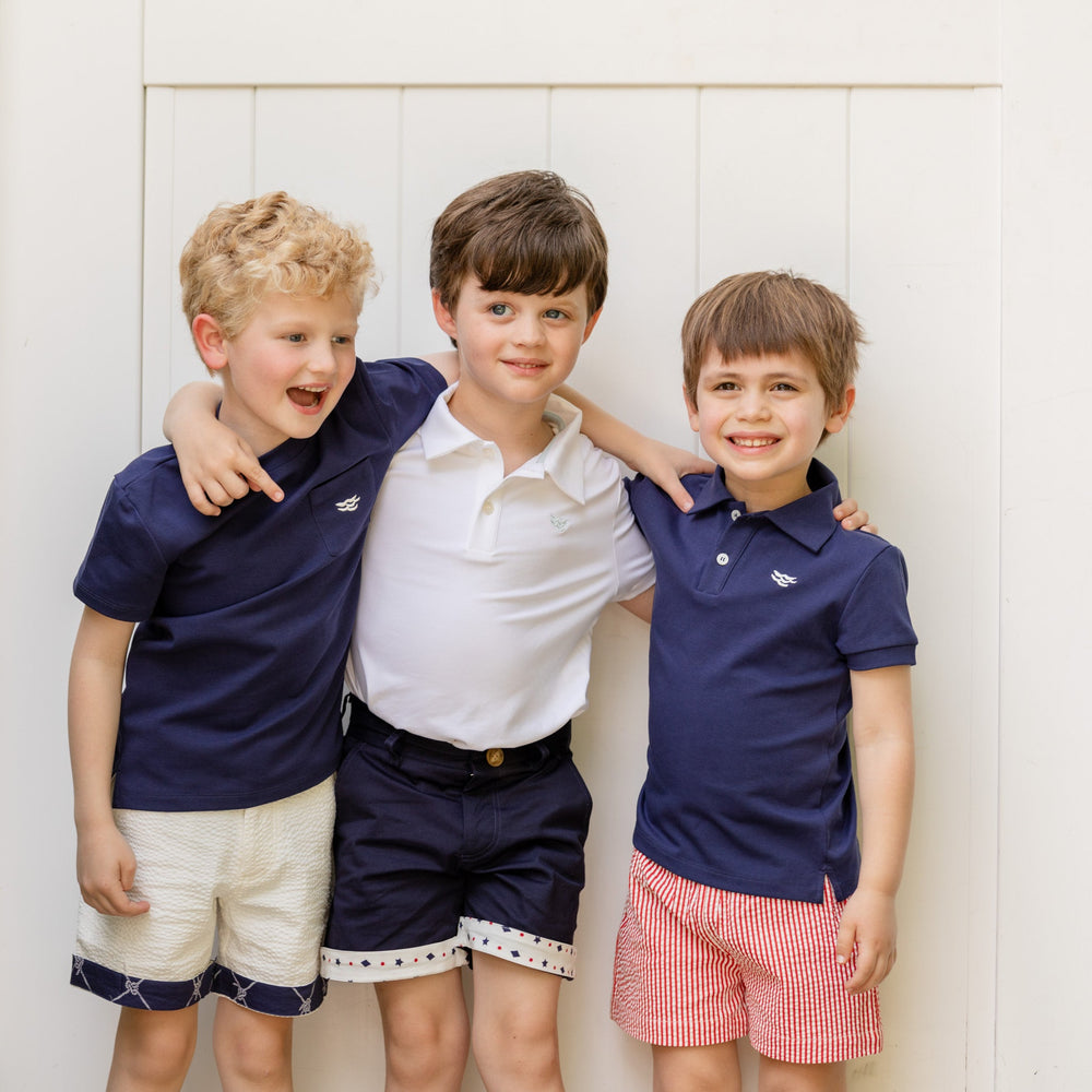 Three boys smiling with arms around each other wearing Carter short-sleeve polos, two in Narragansett Navy and one white against a light paneled wall