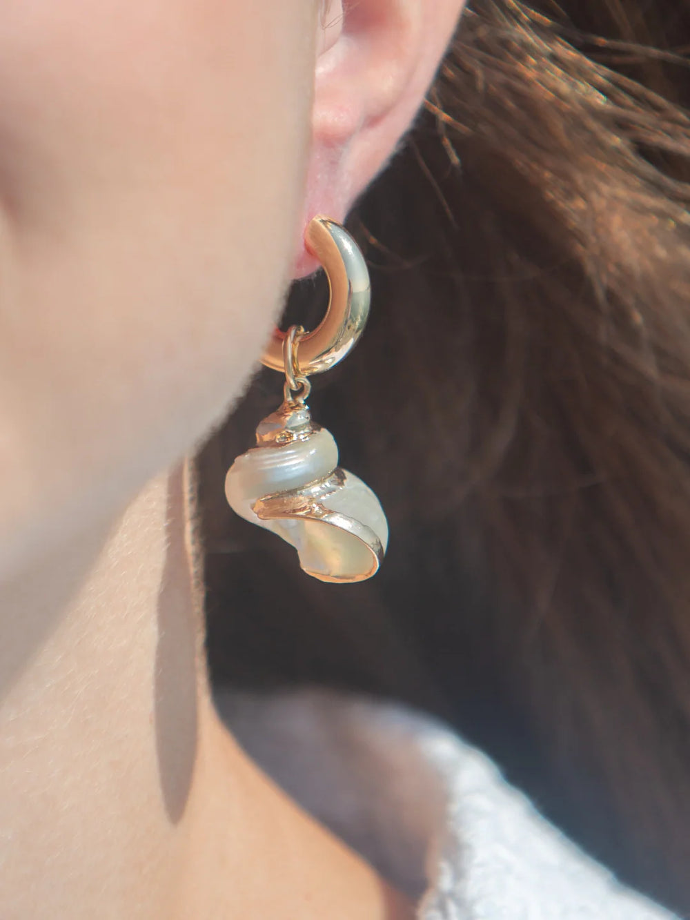 Close-up of Turbo Shell Charm in White on ear showcasing gold-plated spiral shell hanging from a small hoop against a blurred background