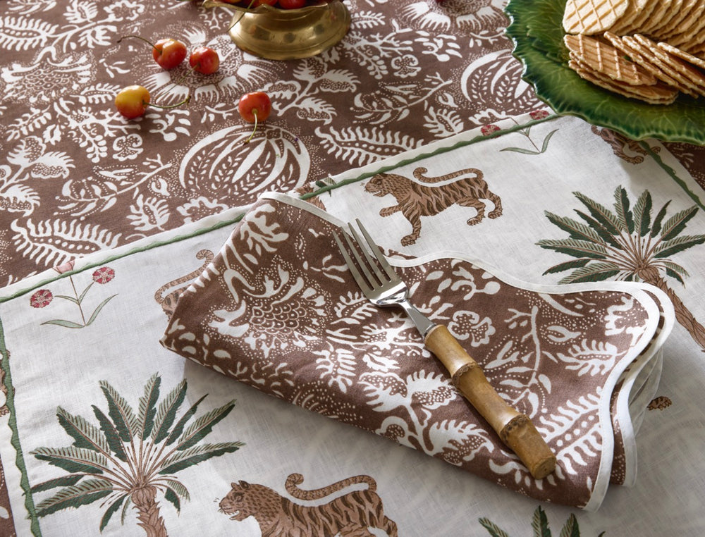 Folded chestnut Granada linen napkin showing white botanical print and scallop-piped edge on tiger-and-palm placemat