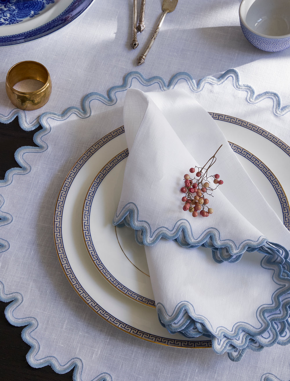 Top down view of Wave napkin folded on a plate showing scalloped hazy blue embroidered trim on white linen