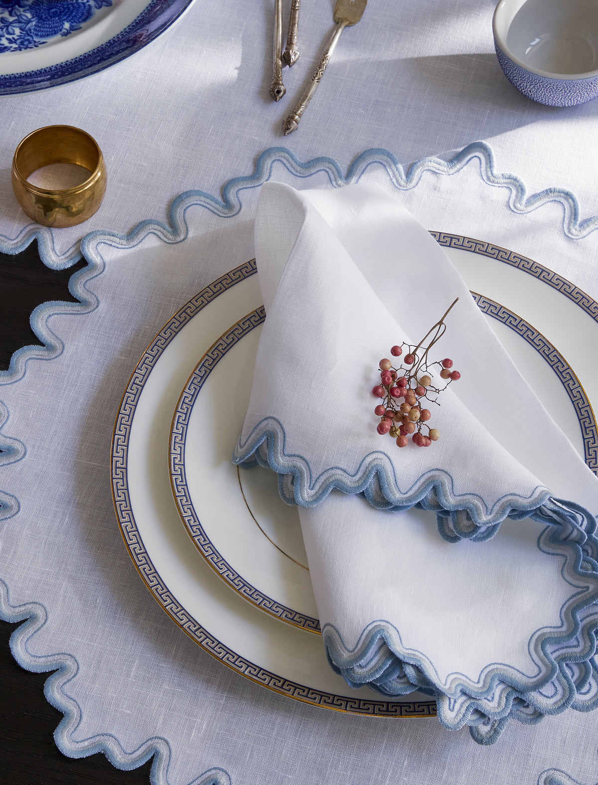 Top down view of Wave napkin folded on a plate showing scalloped hazy blue embroidered trim on white linen