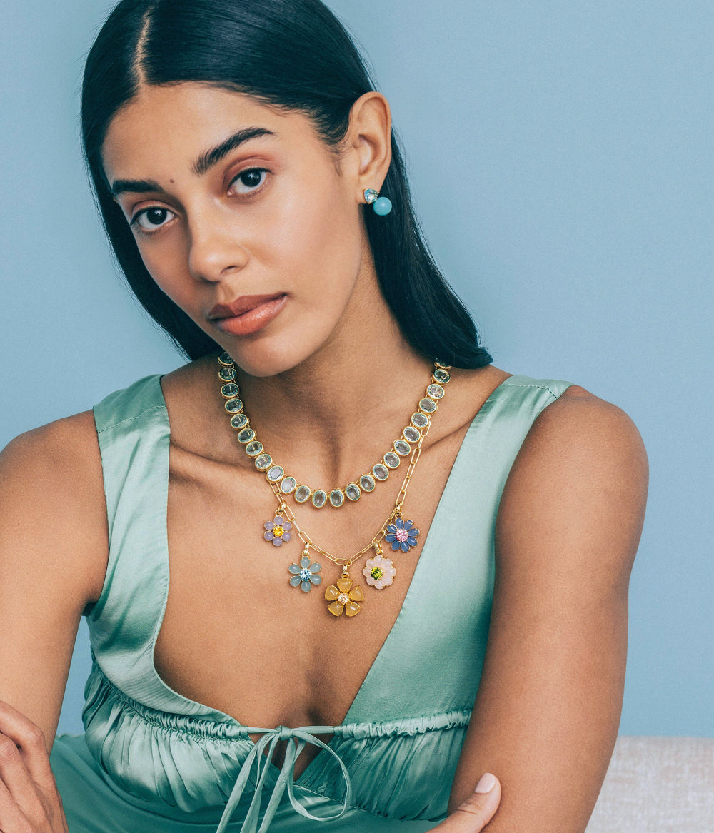 Model wearing Talia Charm Necklace showcasing six pastel flower charms on a gold paperclip chain against blue background