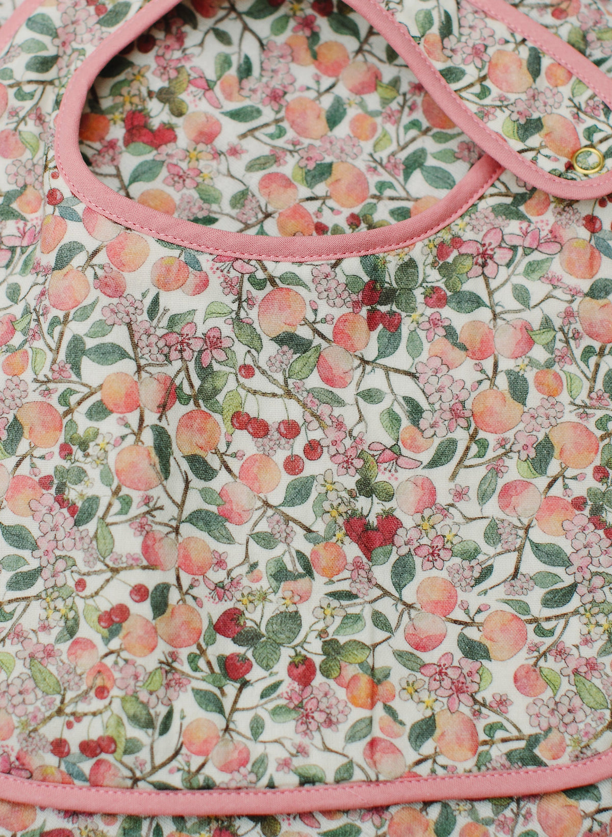 Close up front view of Peach Blossom Bib showcasing its peach blossom print, pink trim, and adjustable snap closure on floral fabric