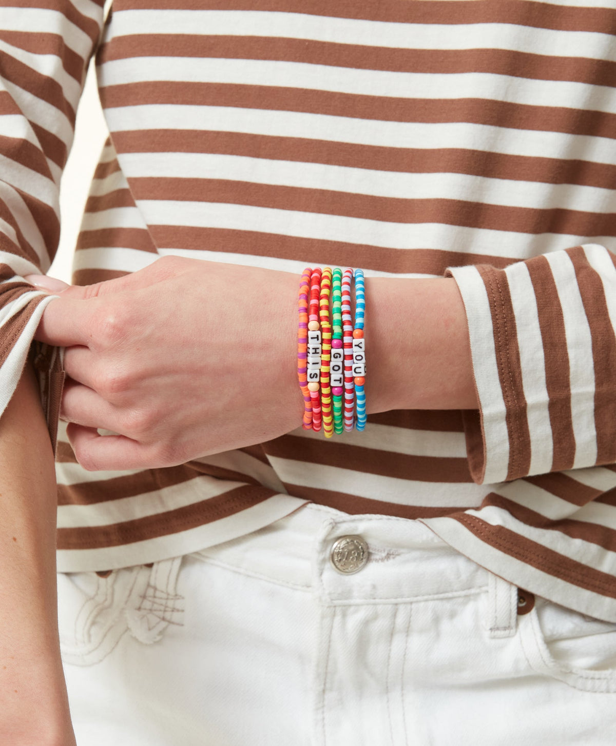 Close-up of wrist wearing five Technicolor Camp beaded bracelets with letter beads reading YOU GOT THIS on a striped top