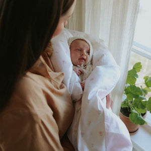 Woman holding a baby wrapped in a white blanket with pink florals by a window with plants.
