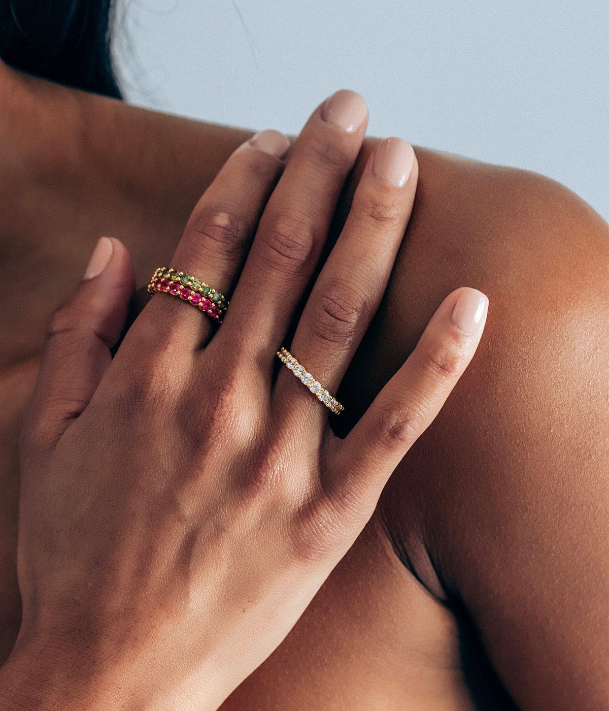 Close-up of hand wearing Theo Ring in Olivine, slim gold-finish eternity band set with green CZ stones against a bare shoulder