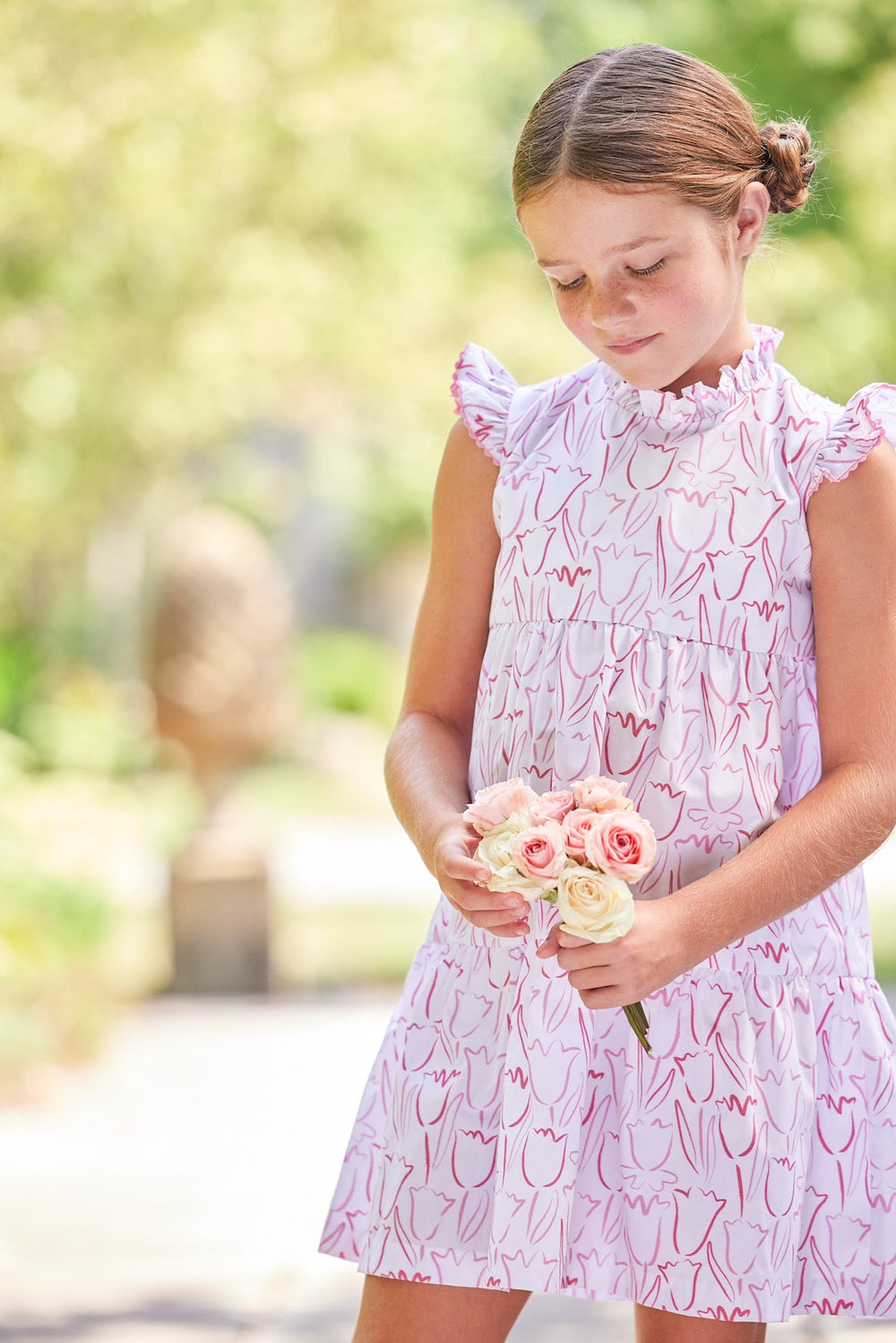 Front view of Tiered Charleston dress in Albany Floral on a girl, ruffled high neck, flutter sleeves and tiered skirt, holding a small bouquet in a garden