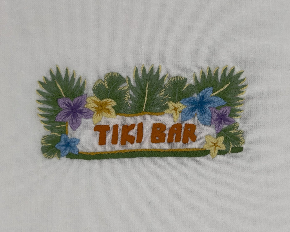 Close up of Tiki Bar tissue box cover showing hand embroidered TIKI BAR with tropical leaves and flowers on white cotton