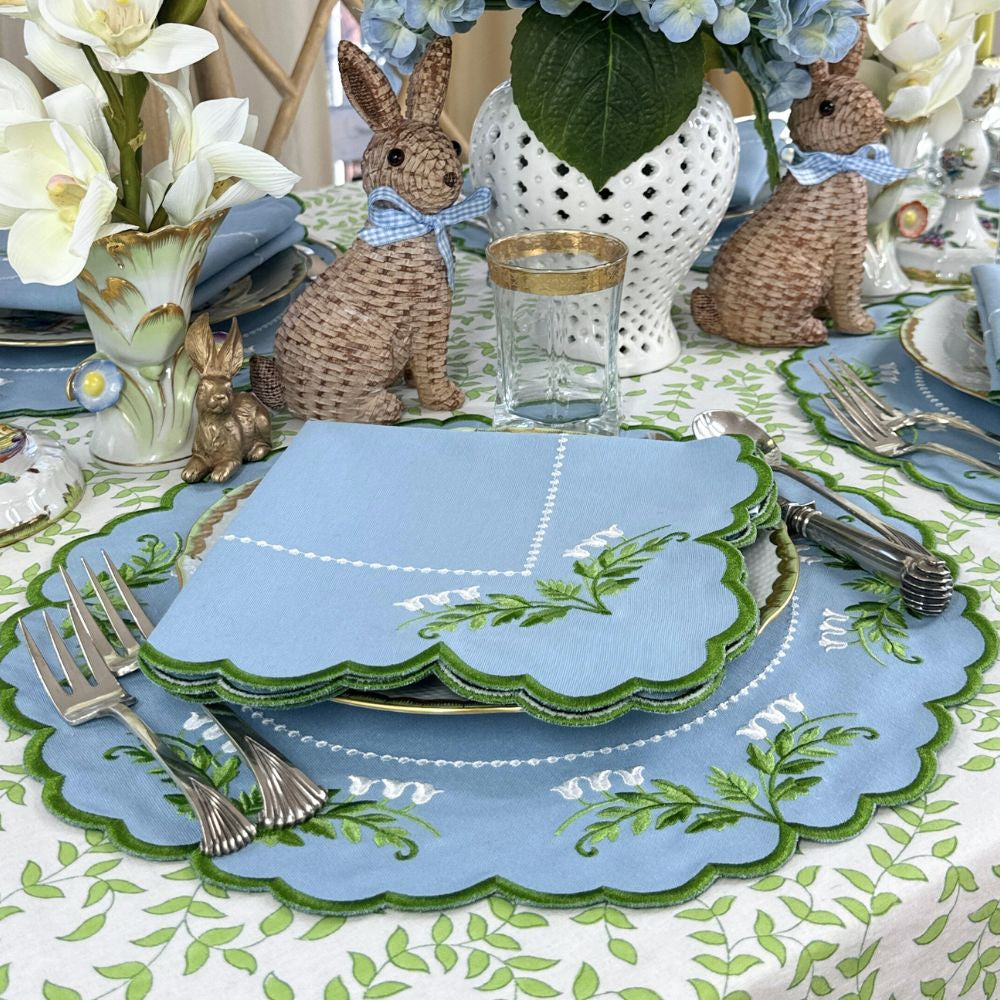 Close-up of Tilly placemat and napkin with blue lily embroidery and scalloped green trim on leaf-print tablecloth