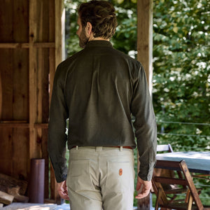 A man outdoors, wearing the Tom Beckbe Dixon Brushed Cotton Twill Shirt and light pants, near wood furniture and greenery.