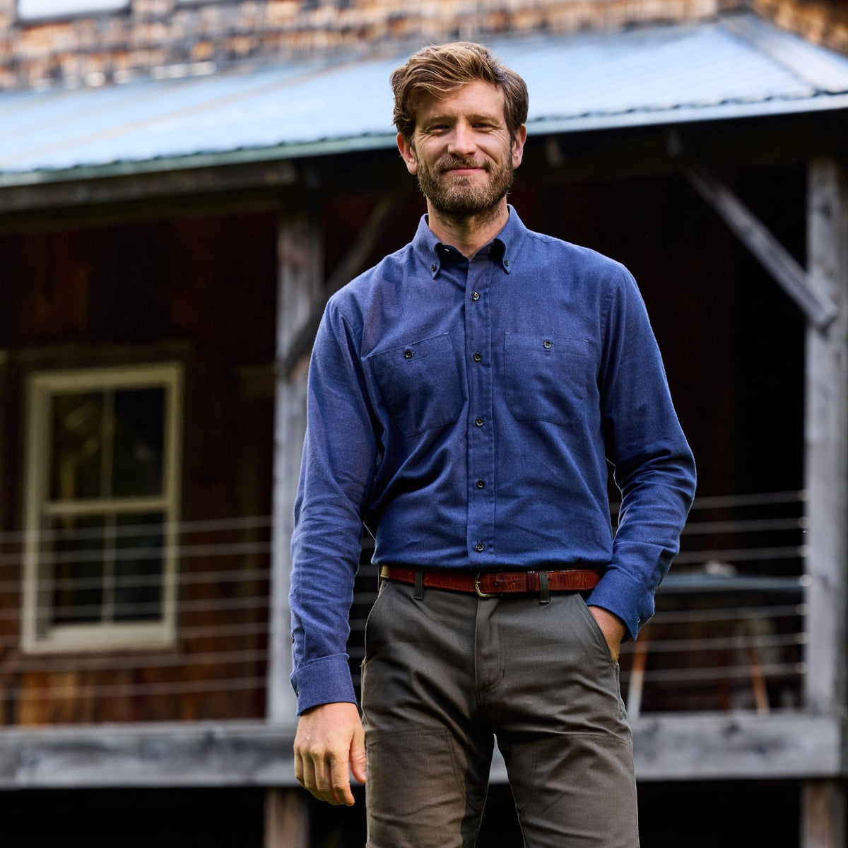 Man wearing a Tom Beckbe Dixon Brushed Cotton Twill Shirt and khaki pants stands outside a rustic wooden building.