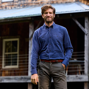 Man wearing a Tom Beckbe Dixon Brushed Cotton Twill Shirt and khaki pants stands outside a rustic wooden building.