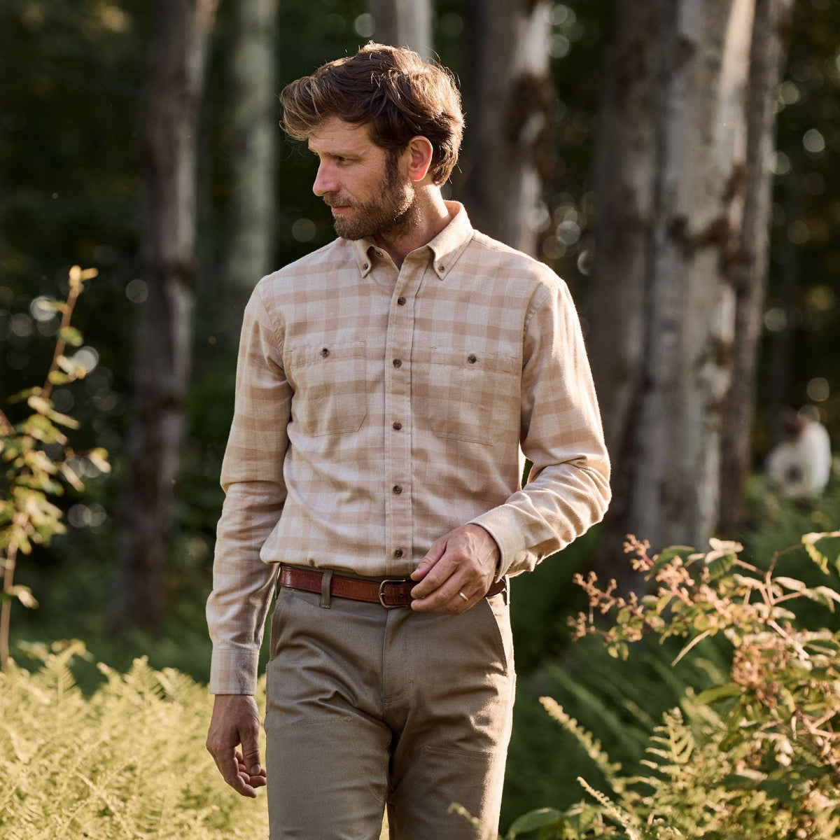 A man in a Tom Beckbe Dixon Brushed Cotton Twill Shirt and khaki pants walks outdoors among tall trees and greenery.
