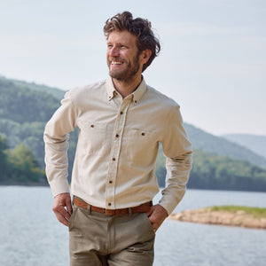 A man in a Tom Beckbe Dixon Brushed Cotton Twill Shirt and khakis smiles by a lake with hills and trees behind him.