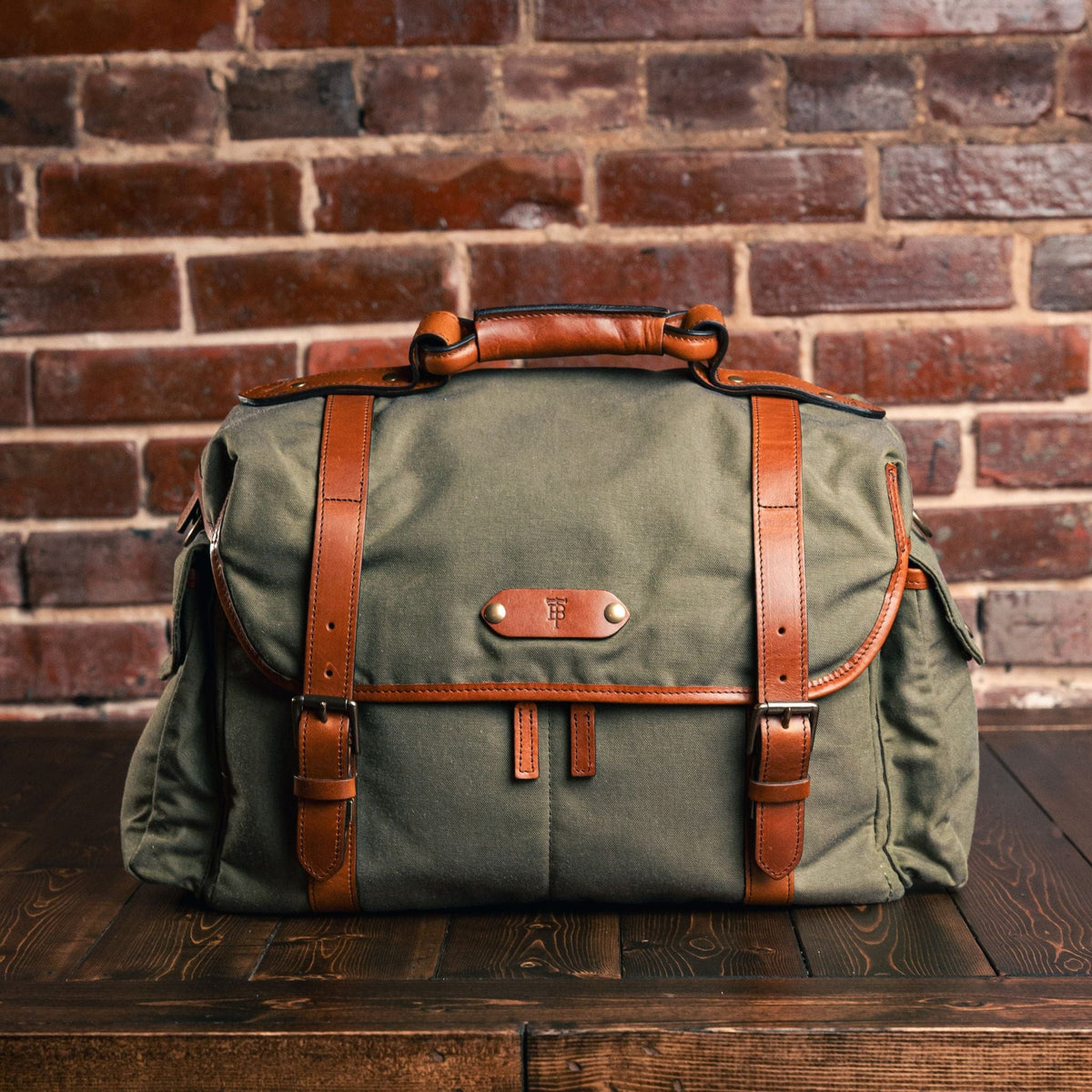 Front view of Sawgrass Canvas Field Bag showcasing its top handle and leather straps on a wooden table against a brick wall