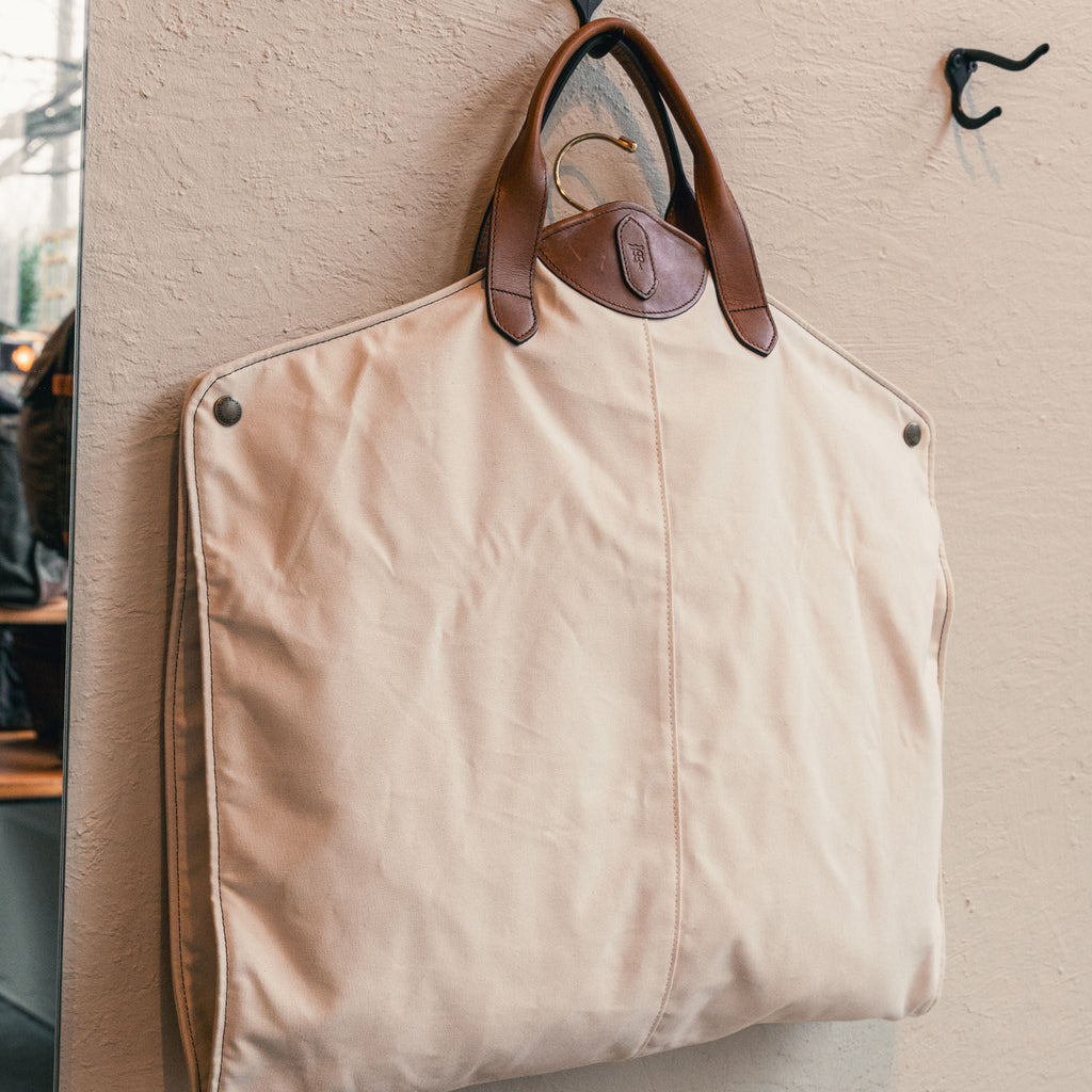 The Perdido Canvas Garment Bag by Tom Beckbe features brown leather handles and hangs from a wall-mounted hook.