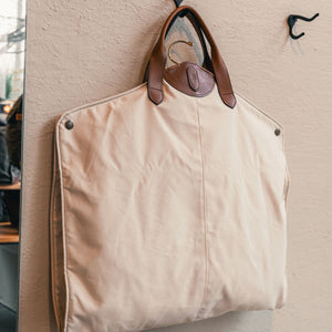 The Perdido Canvas Garment Bag by Tom Beckbe features brown leather handles and hangs from a wall-mounted hook.