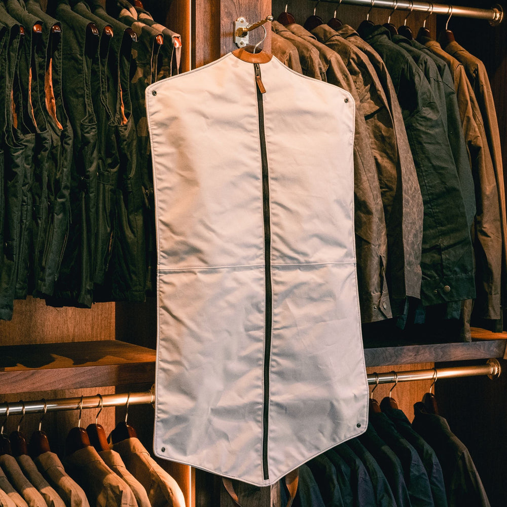 The Tom Beckbe Perdido Canvas Garment Bag in white with a central zipper hangs in front of jackets in a store.