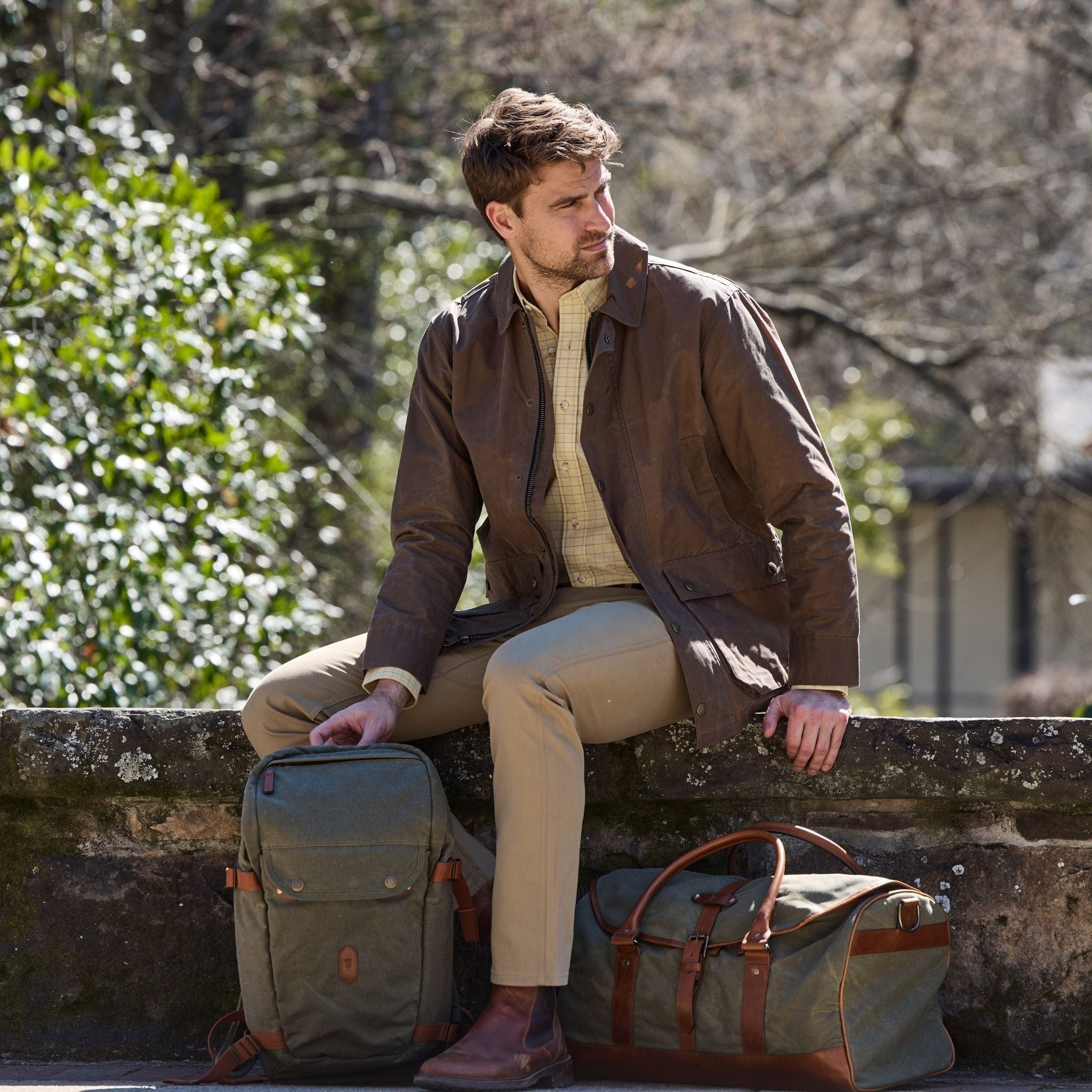 A man sits on a ledge in a brown jacket, beige pants, with two Tom Beckbe Canvas Weekender Duffel Bags beside him.