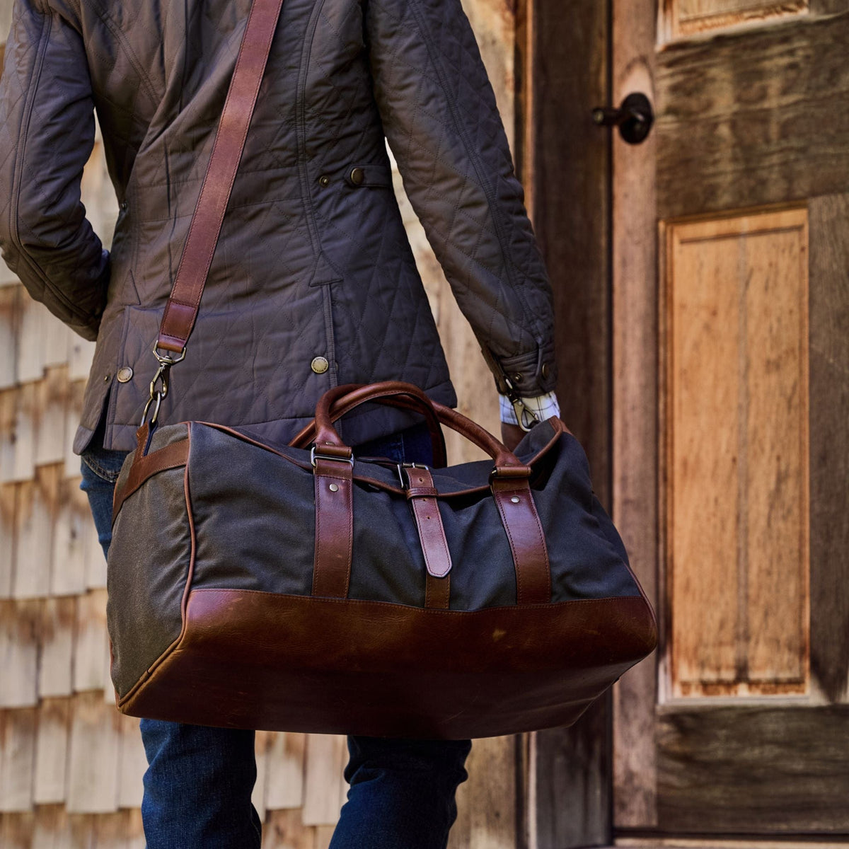 A travel companion in a dark jacket with a Tom Beckbe Canvas Weekender Duffel Bag approaches a wooden door.