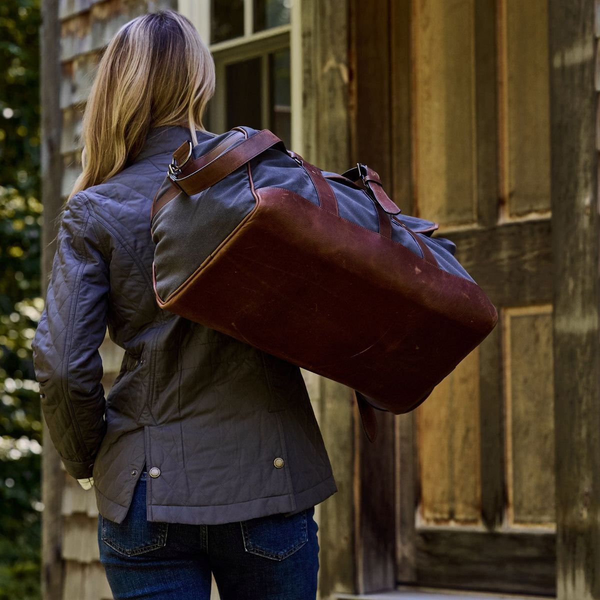 A person with long hair and a Tom Beckbe Canvas Weekender Duffel stands before a wooden building.