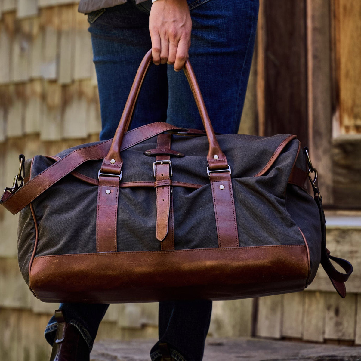 Person holding a Canvas Weekender Duffel Bag by Tom Beckbe, featuring brown leather accents, in front of a wooden building.