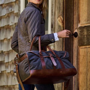 Individual in a brown quilted jacket with a Tom Beckbe Canvas Weekender Duffel Bag, opening a wooden door.