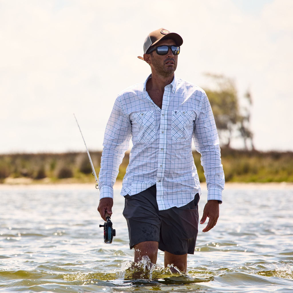 A man in Tom Beckbes Crenshaw Performance Shorts wades through waist-deep water, fishing under a partly cloudy sky.